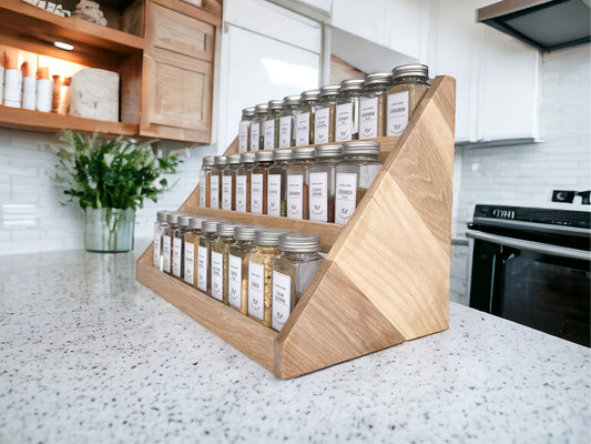 A three-tiered wooden display holding herb and spice jars sitting on a granite countertop in a kitchen.